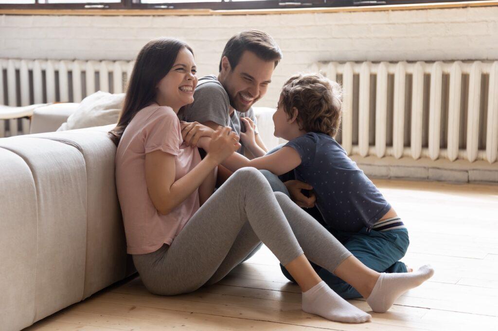 Eine fröhliche Familie sitzt lachend auf dem Boden vor einem Heizkörper, während sie Zeit zusammen verbringt.